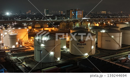 High-angle night view of a fuel storage tank terminal with multiple large, illuminated cylinders and a complex piping infrastructure at an industrial port facility, glowing in the dark. 139244694