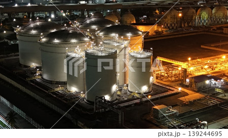 High-angle night view of a fuel storage tank terminal with multiple large, illuminated cylinders and a complex piping infrastructure at an industrial port facility, glowing in the dark. High-angle night view of a fuel storage tank terminal with multiple large, illuminated cylinders and a complex piping infrastructure at an industrial port facility, glowing in the dark. 139244695