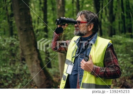Far away, looking through the binoculars. Senior forest ecologist is working in the woods. In the uniform Far away, looking through the binoculars. Senior forest ecologist is working in the woods. In the uniform 139245253