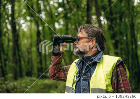 Professional binoculars in hands. Senior forest ecologist is working in the woods. In the uniform Professional binoculars in hands. Senior forest ecologist is working in the woods. In the uniform 139245260