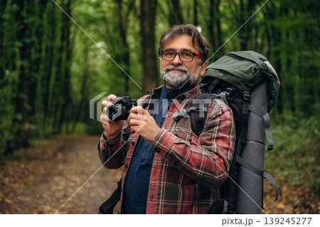 Camera is in hands, making photos. Hiking experience, senior man is in the forest at daytime 139245277