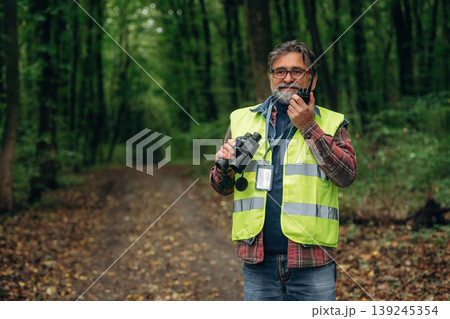 Radio walkie talkie and binoculars in hands. Senior forest ecologist is working in the woods. In the uniform 139245354