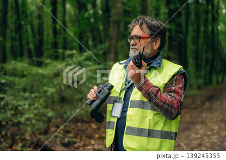Radio walkie talkie and binoculars in hands. Senior forest ecologist is working in the woods. In the uniform 139245355
