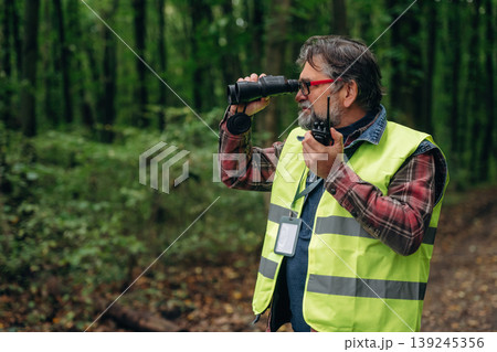 Radio walkie talkie and binoculars in hands. Senior forest ecologist is working in the woods. In the uniform 139245356