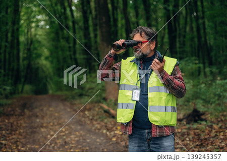 Radio walkie talkie and binoculars in hands. Senior forest ecologist is working in the woods. In the uniform 139245357