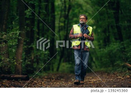 Binoculars in hands, walking. Senior forest ecologist is working in the woods. In the uniform Binoculars in hands, walking. Senior forest ecologist is working in the woods. In the uniform 139245360