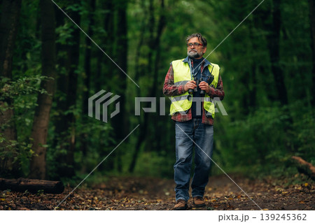 Binoculars in hands, walking. Senior forest ecologist is working in the woods. In the uniform Binoculars in hands, walking. Senior forest ecologist is working in the woods. In the uniform 139245362