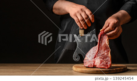 Chef cutting raw t-bone steak with cleaver on wooden board, dark background. copy space Chef cutting raw t-bone steak with cleaver on wooden board, dark background. copy space 139245454