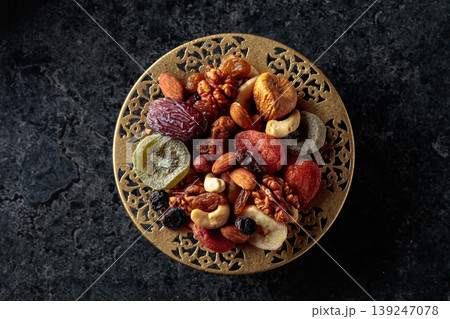 Dried tropical fruits, nuts, and raisins in a golden bowl on black stone table. Dried tropical fruits, nuts, and raisins in a golden bowl on black stone table. 139247078
