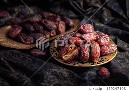 Dates fruits in a golden bowl on black fabric with gold ornamentation. Dates fruits in a golden bowl on black fabric with gold ornamentation. 139247108