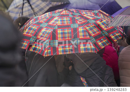 Plaid umbrella with raindrops covers people in crowd during rainy city event. Concept of umbrella protection, weather, travel, commuting, seasonal rain, street life, public gathering Plaid umbrella with raindrops covers people in crowd during rainy city event. Concept of umbrella protection, weather, travel, commuting, seasonal rain, street life, public gathering 139248283