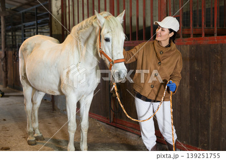 Female stable worker leads a white horse out of stall Female stable worker leads a white horse out of stall 139251755