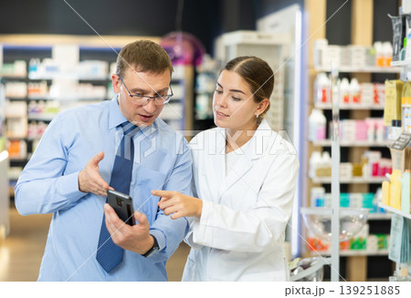 Friendly female pharmacist helping adult man with electronic prescription in phone choosing medical supplies at drugstore 139251885