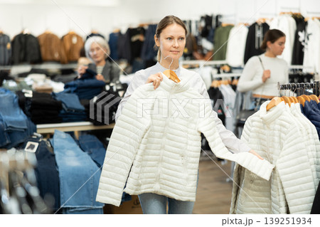 Positive woman chooses white winter down jacket in clothing store Positive woman chooses white winter down jacket in clothing store 139251934