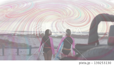 Walking surfers wearing rash guards carrying surfboards on sandy beach, with fence and rear of car 139253130