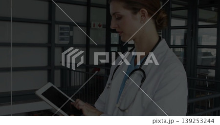 Reviewing doctor in blue scrubs in white coat using tablet on hospital corridor with stethoscope Reviewing doctor in blue scrubs in white coat using tablet on hospital corridor with stethoscope 139253244