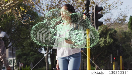 Standing woman holding smartphone at street corner showing holographic cloud with gear icons 139253554