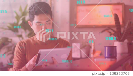 Chinese man wearing brown shirt tapping tablet at desk in home office, floating currency symbols 139253705