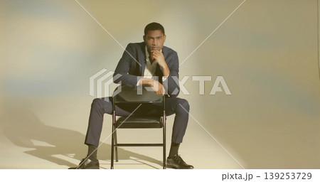 Posing adult man wearing navy suit leaning forward on folding chair in studio, with thoughtful pose Posing adult man wearing navy suit leaning forward on folding chair in studio, with thoughtful pose 139253729
