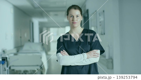 Standing mid adult nurse wearing navy scrubs crossing arms in hospital corridor, with bed rail 139253794