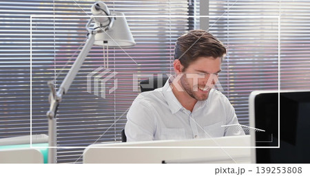 Smiling man typing on keyboard and glancing at monitor in office, with desk lamp and blinds Smiling man typing on keyboard and glancing at monitor in office, with desk lamp and blinds 139253808