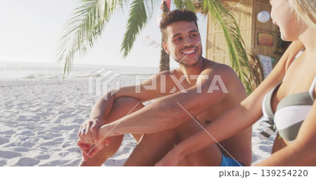 Smiling couple sitting on sand under palm by ocean waves, with thatched-roof hut and paper-lanterns Smiling couple sitting on sand under palm by ocean waves, with thatched-roof hut and paper-lanterns 139254220
