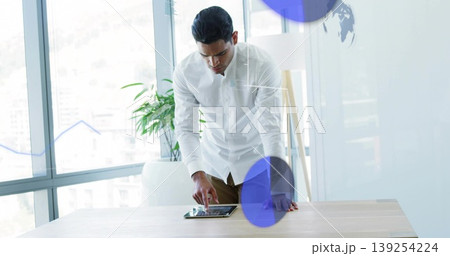 Leaning man in white shirt, brown trousers using tablet at desk in office with data overlays 139254224