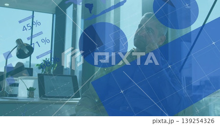 Businessman in suit sitting at desk in office, with laptop and lamp while viewing data graphics Businessman in suit sitting at desk in office, with laptop and lamp while viewing data graphics 139254326