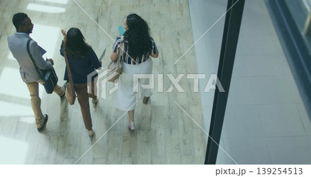 Walking professionals carrying bags and documents through light office corridor in formal wear Walking professionals carrying bags and documents through light office corridor in formal wear 139254513