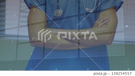 Standing male doctor crossing arms in corridor wearing scrubs, stethoscope with digital interface Standing male doctor crossing arms in corridor wearing scrubs, stethoscope with digital interface 139254557