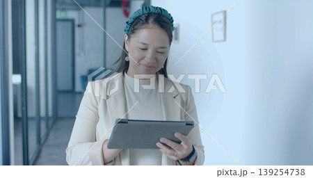 Standing mid adult Asian woman wearing beige blazer holding tablet in office corridor Standing mid adult Asian woman wearing beige blazer holding tablet in office corridor 139254738