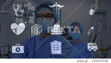 Crossing arms male surgeon in blue scrubs in operating room, with overhead lights and medical icons Crossing arms male surgeon in blue scrubs in operating room, with overhead lights and medical icons 139254780