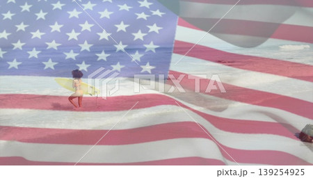 Walking surfer wearing swimwear, carrying yellow surfboard on shallow shore, with US flag overlay 139254925