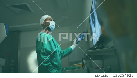 Reviewing surgeon in green scrubs consulting wall-mounted scans in operating room, with tablet 139254926