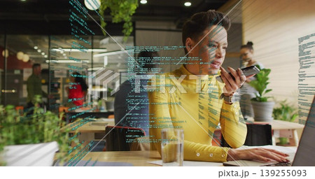 Typing woman in yellow sweater talking into smartphone at office with code on glass partition 139255093