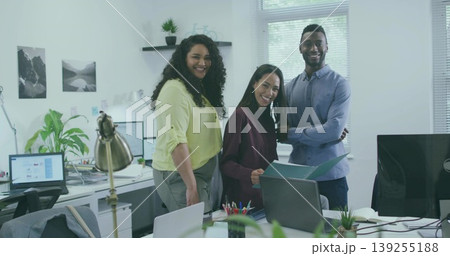 Smiling three coworkers wearing work attire standing at workstation in office, with laptop monitors Smiling three coworkers wearing work attire standing at workstation in office, with laptop monitors 139255188