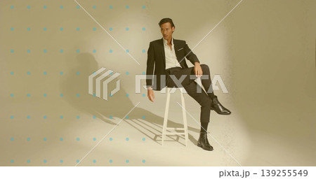 Asian male model wearing black suit posing on stool in studio, with blue dot grid backdrop 139255549