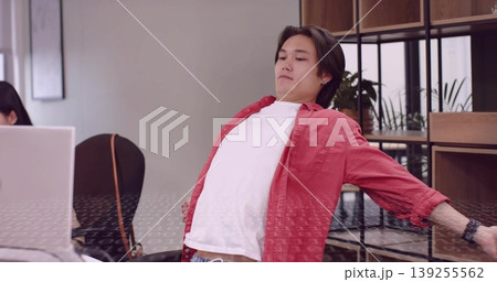 Stretching coworker in red shirt leaning back at office desk, with dual laptops and plant shelves Stretching coworker in red shirt leaning back at office desk, with dual laptops and plant shelves 139255562