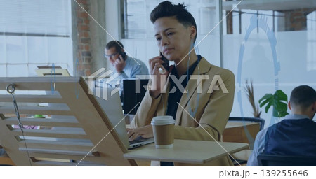 Holding smartphone, Asian woman in blazer typing on laptop at desk in office, with coffee cup Holding smartphone, Asian woman in blazer typing on laptop at desk in office, with coffee cup 139255646