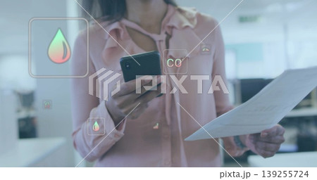 Female office worker analyzing overlay in open-plan office, with smartphone, document and CO2 icons 139255724