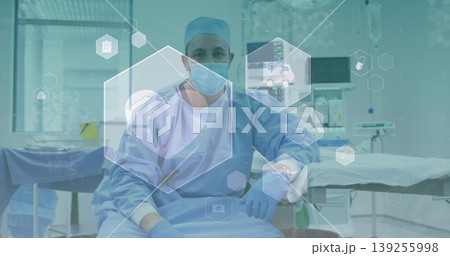 Sitting male surgeon wearing blue scrubs with mask and gloves in surgical suite with monitors Sitting male surgeon wearing blue scrubs with mask and gloves in surgical suite with monitors 139255998