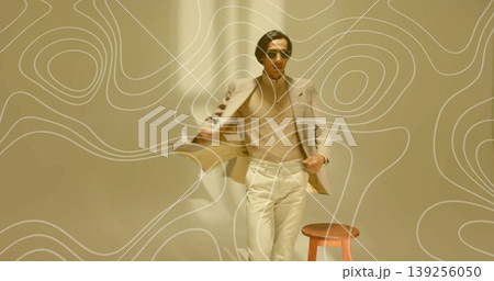 Standing Asian male model wearing beige blazer and trousers in minimalist studio, with wooden stool Standing Asian male model wearing beige blazer and trousers in minimalist studio, with wooden stool 139256050