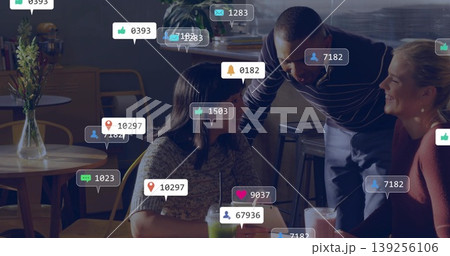 Chatting friends leaning at coffee shop wooden table sipping drinks with floating social icons Chatting friends leaning at coffee shop wooden table sipping drinks with floating social icons 139256106