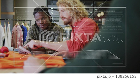 Reviewing designers analyzing garments in studio with tablet, orange fabric and digital overlays 139256155
