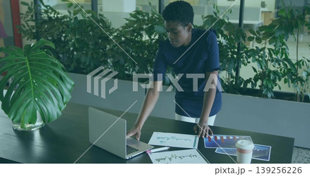 Businesswoman wearing business attire using laptop and charts at office table, with potted plant Businesswoman wearing business attire using laptop and charts at office table, with potted plant 139256226