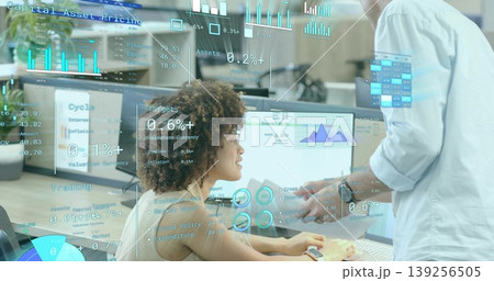 Seated woman and standing coworker reviewing financial charts on monitors in office, with documents Seated woman and standing coworker reviewing financial charts on monitors in office, with documents 139256505