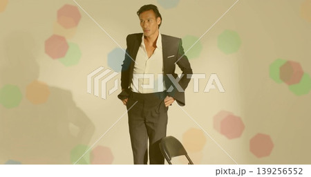 Standing Asian man wearing suit behind black metal folding chair in studio with hexagon projections 139256552