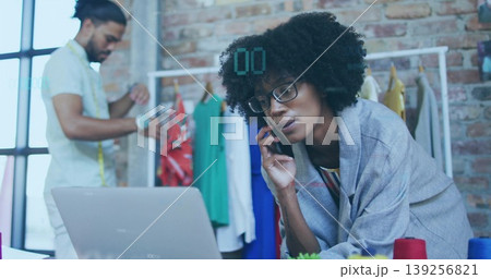 Female designer in shawl leaning over laptop, speaking on smartphone in studio with measuring tape 139256821