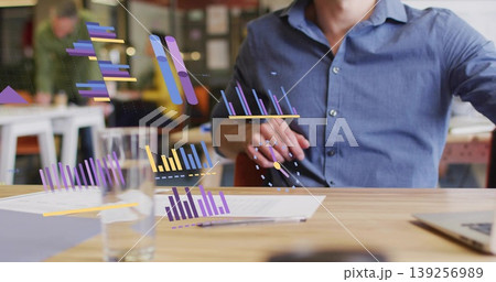 Reviewing man in blue shirt manipulating digital bar charts at office desk, with printed documents 139256989