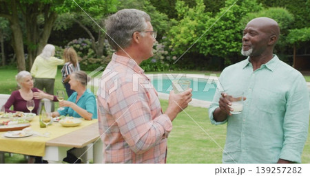 Holding glasses, two men conversing in backyard garden beside pool, with wooden table yellow cloth 139257282